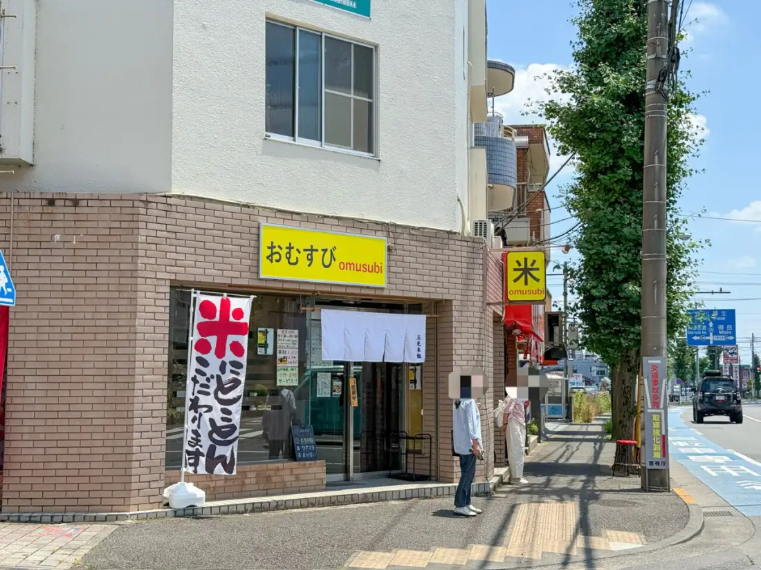 三光米飯の外観、日中の店頭。レンガ壁に黄色の横看板『おむすび omusubi』、白暖簾『三光米飯』、赤の縦看板『米』が並ぶ。