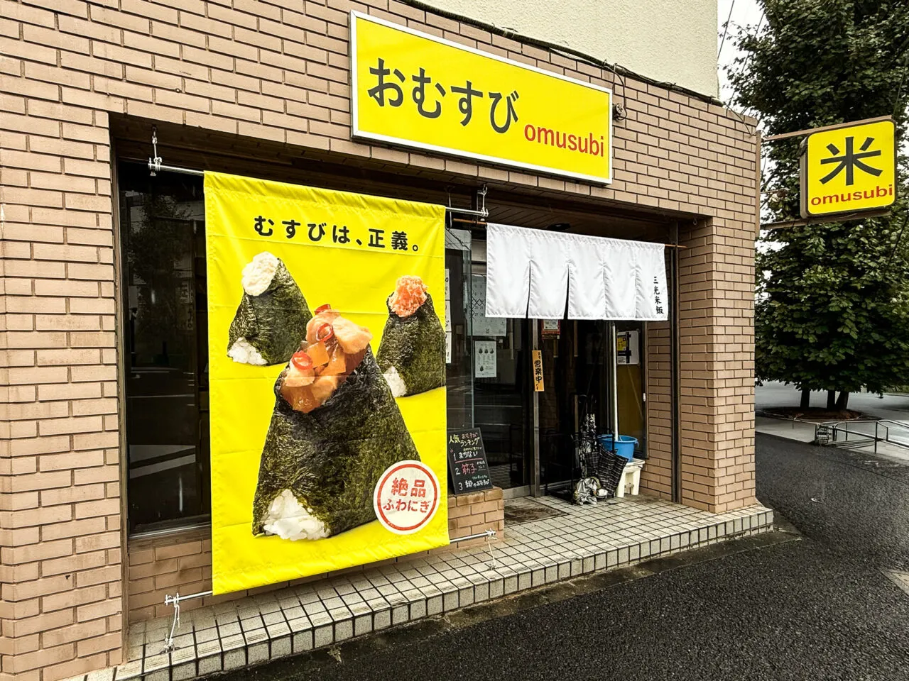 三光米飯の店頭。黄色い垂れ幕に『むすびは、正義。』のキャッチコピーとおむすびのイラストが描かれ、上に黄色の横看板『おむすび omusubi』、右手に赤の縦看板『米』が並ぶ、レンガ調のファサード。