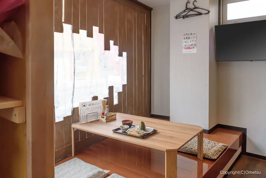 座敷の内装。縦格子の茶色木壁と市松柄の座布団、ライトウッドの低座テーブル。