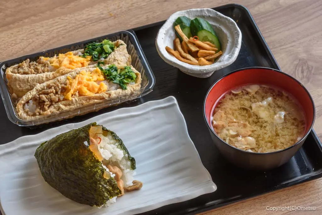 定食のセット。三光いなりとおむすび、味噌汁とお新香が黒盆に並ぶ。